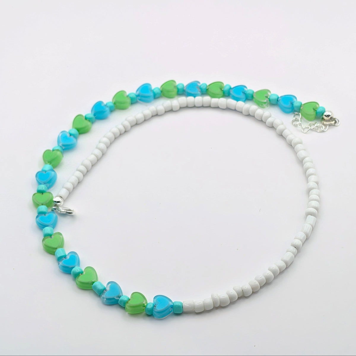 A beaded necklace featuring alternating strands of white round beads, light blue heart-shaped beads, and lime green heart-shaped beads, displayed on a clean white background.