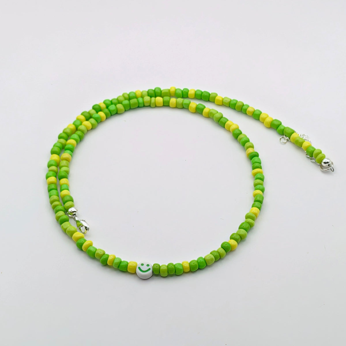 A beaded necklace made of alternating green and yellow round beads, featuring a small silver smiley face charm in the center and silver metal clasp ends, displayed on a clean white background.