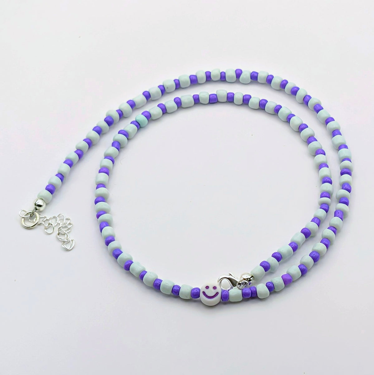 A beaded necklace with alternating light blue-gray and purple beads, featuring a small white smiley face charm and silver-tone lobster clasp on a plain white background.