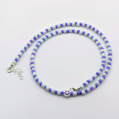 A beaded necklace with alternating light blue-gray and purple beads, featuring a small white smiley face charm and silver-tone lobster clasp on a plain white background.