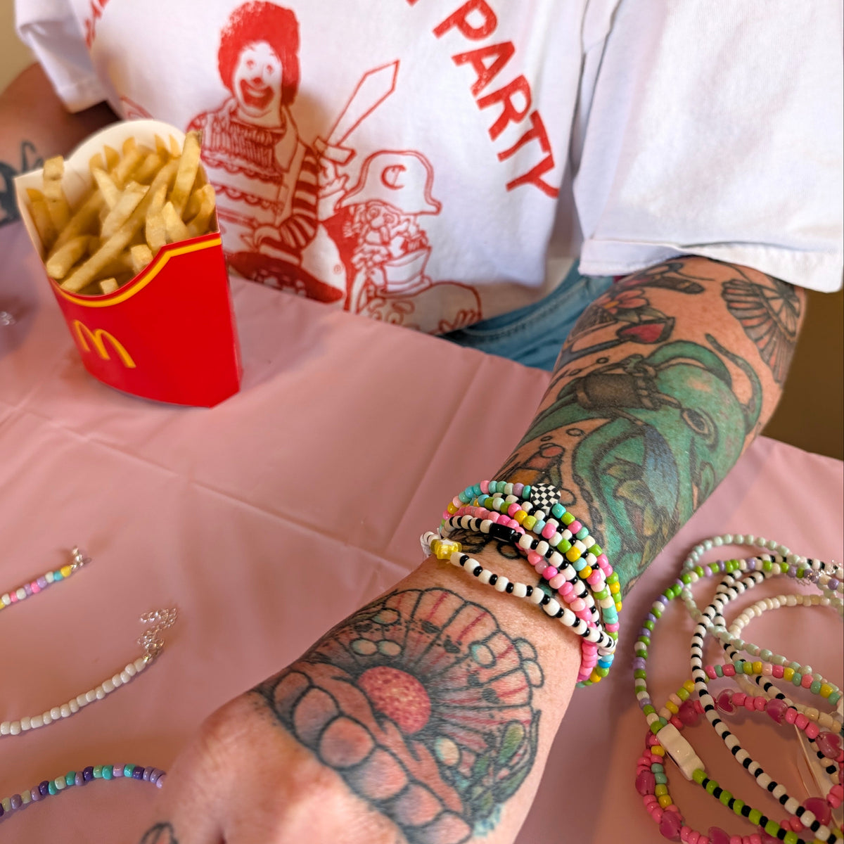 A close-up of a person's tattooed forearm wearing multiple colorful beaded bracelets, with more bracelets on the table and a red McDonald's fries container nearby. The person is wearing a white graphic T-shirt.