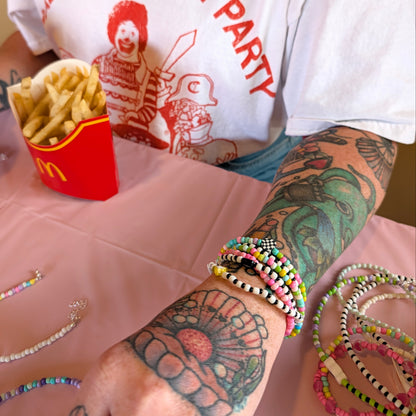 A close-up of a person's tattooed forearm wearing multiple colorful beaded bracelets, with more bracelets on the table and a red McDonald's fries container nearby. The person is wearing a white graphic T-shirt.