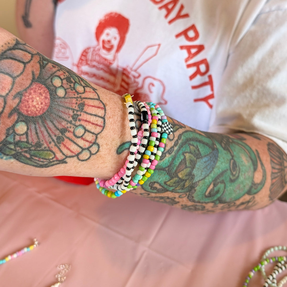 A close-up of a person's tattooed arm wearing multiple colorful beaded bracelets, with a white graphic T-shirt and a pink tablecloth visible in the background.