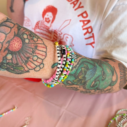 A close-up of a person's tattooed arm wearing multiple colorful beaded bracelets, with a white graphic T-shirt and a pink tablecloth visible in the background.