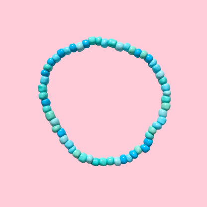 A beaded stretch bracelet made of small cylindrical beads in a gradient of turquoise, blue, and teal, displayed on a textured white background.