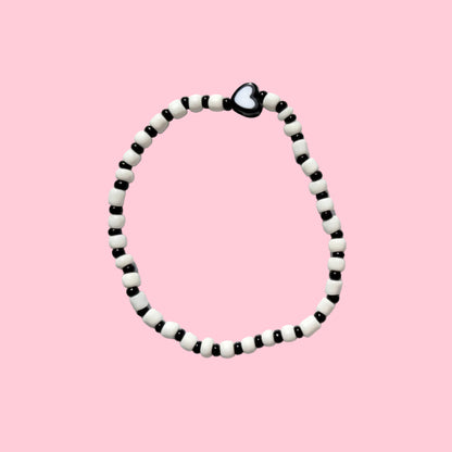 A beaded necklace with alternating black and white beads, featuring a small black heart charm at the top, displayed against a textured white wall.