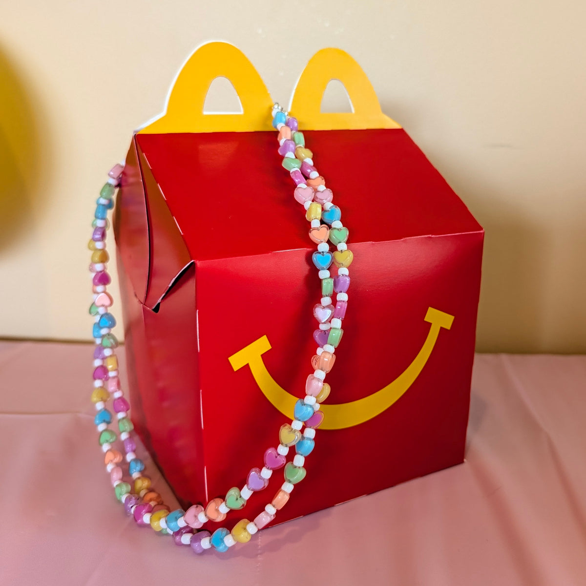 A red McDonald's Happy Meal box with yellow handle cutouts and smiley face detail, decorated with a pastel beaded necklace, set on a pink tablecloth with a birthday banner in the background.