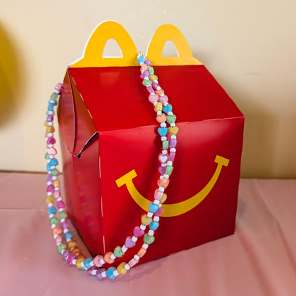 A red McDonald's Happy Meal box with yellow handle cutouts and smiley face detail, decorated with a pastel beaded necklace, set on a pink tablecloth with a birthday banner in the background.