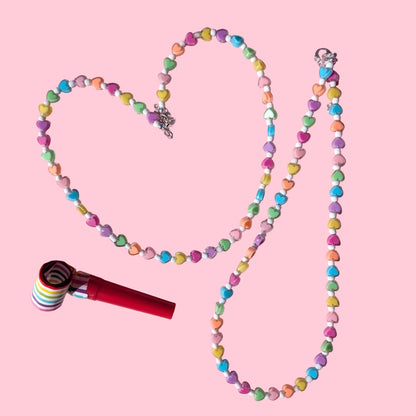 A beaded necklace made of pastel candy-coated beads arranged in a heart shape on a pink background, with a red and white striped lollipop-style charm placed beside it.
