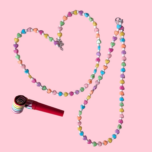 A beaded necklace made of pastel candy-coated beads arranged in a heart shape on a pink background, with a red and white striped lollipop-style charm placed beside it.