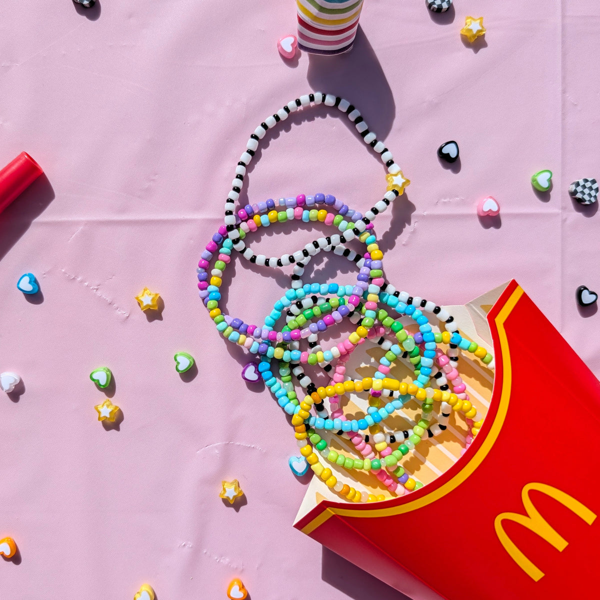 A styled flat-lay featuring colorful beaded necklaces and bracelets spilling out of a red McDonald's french fries box on a pink background, alongside a red party horn and striped party hats.