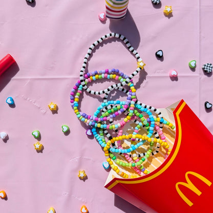 A styled flat-lay featuring colorful beaded necklaces and bracelets spilling out of a red McDonald's french fries box on a pink background, alongside a red party horn and striped party hats.