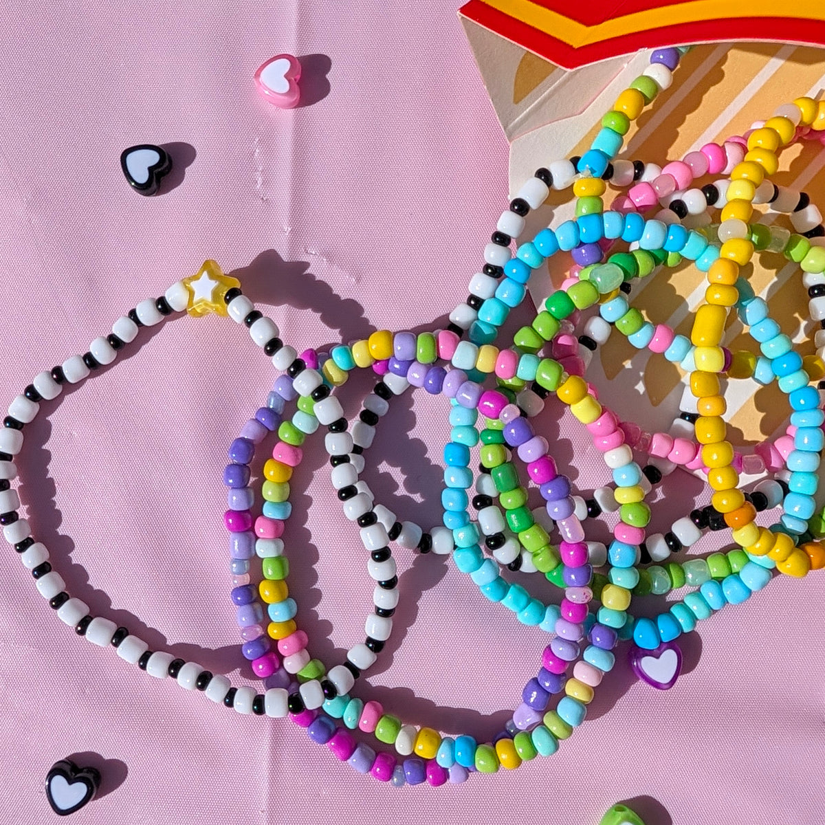 A collection of colorful beaded bracelets and necklaces spilled from a red and yellow box onto a pink background, surrounded by small heart-shaped charms.