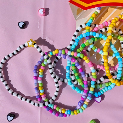A collection of colorful beaded bracelets and necklaces spilled from a red and yellow box onto a pink background, surrounded by small heart-shaped charms.