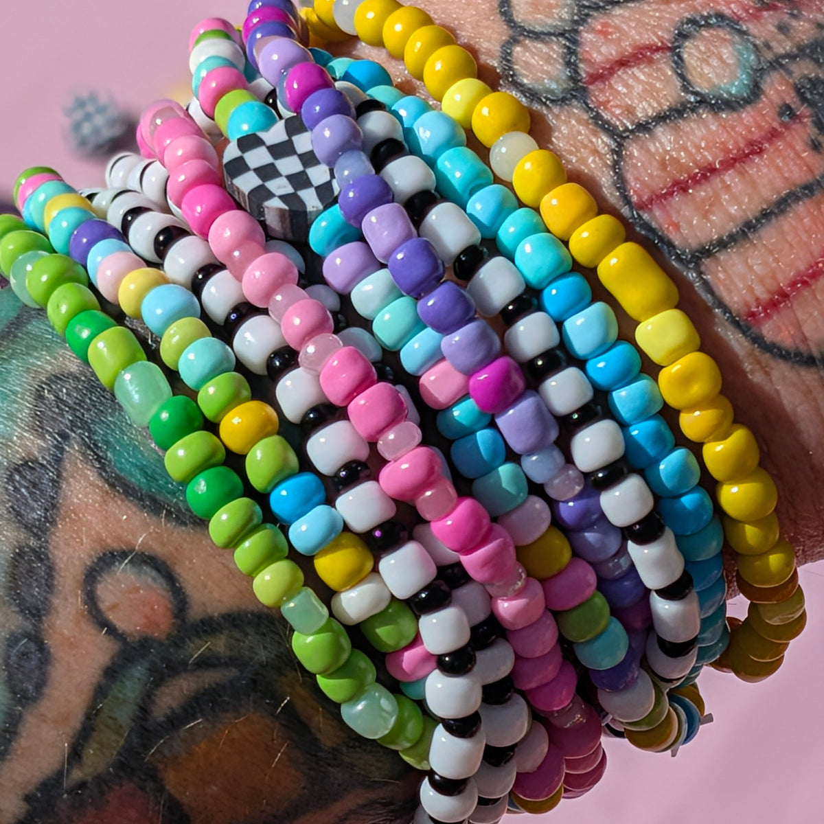 A close-up of a tattooed wrist wearing a stack of colorful beaded stretch bracelets in yellow, blue, pink, green, purple, black, and white, set against a solid pink background.