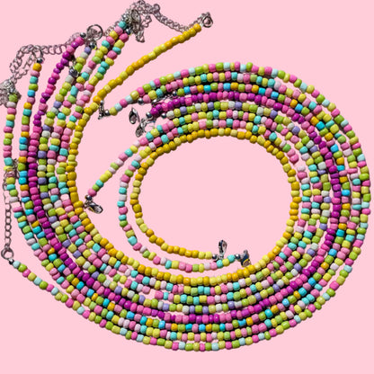 A multi-strand beaded necklace arranged in a spiral on a light pink background. The necklace features small, round beads in pastel pink, yellow, green, and blue, with silver-tone metal chains and clasps.