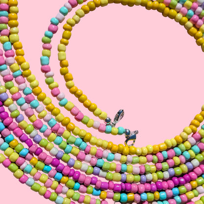 A close-up of multiple strands of pastel-colored cylindrical beads arranged in loops on a pink background. The beads come in pink, yellow, mint green, light blue, and lime green, with small silver metal clasps visible.