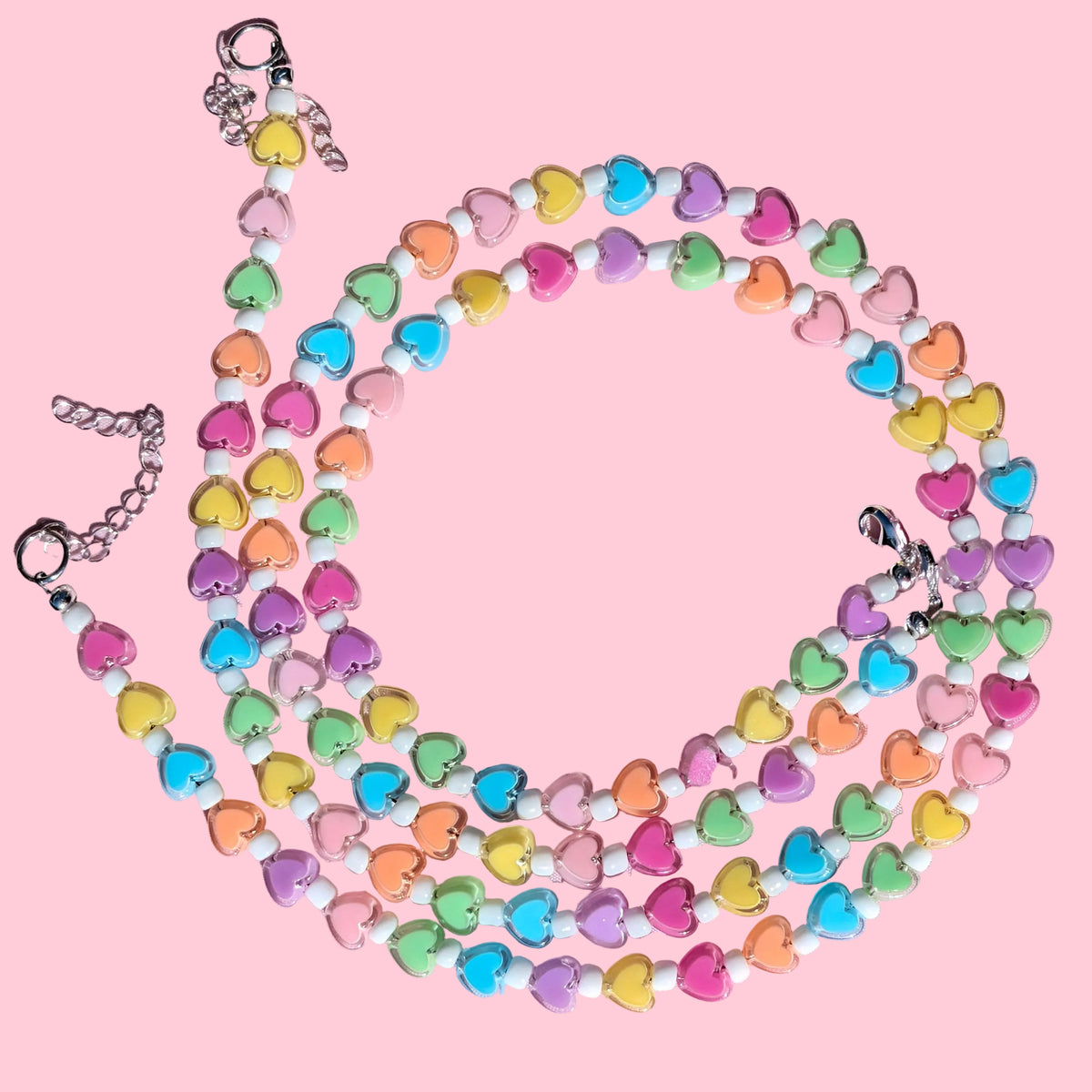 A multi-strand beaded necklace made of pastel heart-shaped beads in pink, yellow, green, blue, orange, and purple, arranged in a circle on a light pink background.