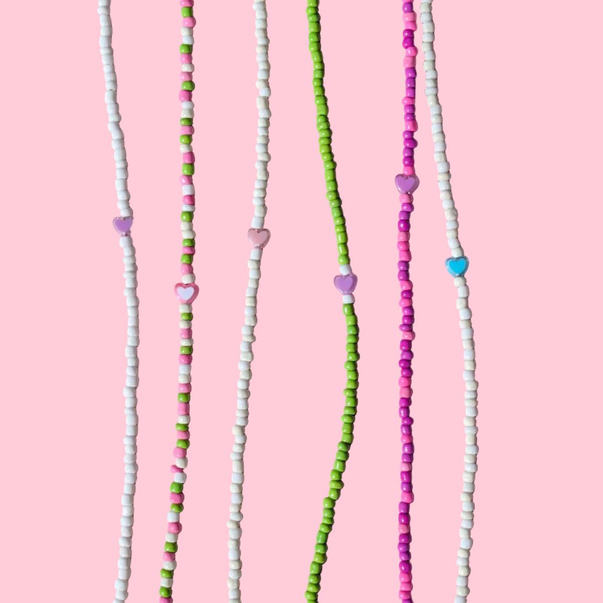 Six beaded strands laid out vertically on a light pink background. The beads are primarily white, green, and pink, with small heart-shaped beads and a few blue accents.