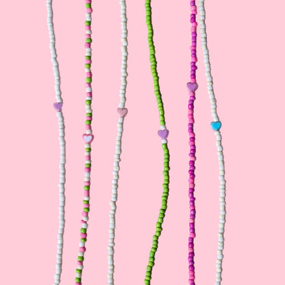 Six beaded strands laid out vertically on a light pink background. The beads are primarily white, green, and pink, with small heart-shaped beads and a few blue accents.