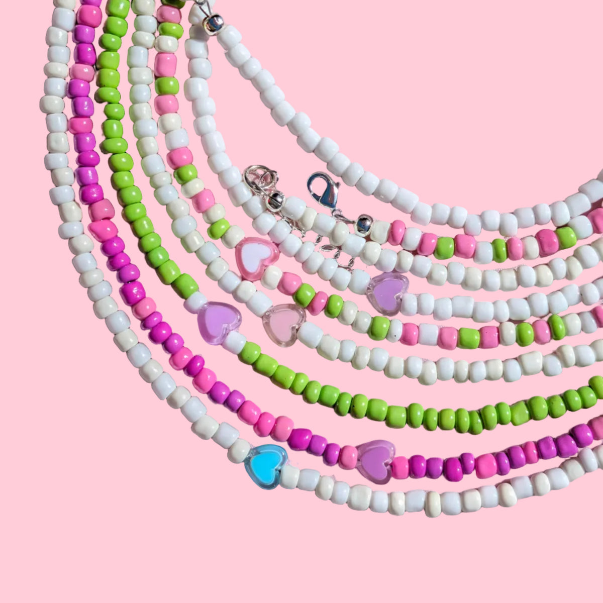 A set of beaded necklaces arranged in overlapping concentric circles on a pink background. The necklaces feature small round beads in white, pink, and lime green, with some pieces including small heart-shaped charms in blue, pink, and purple, and silver-toned metal clasps.