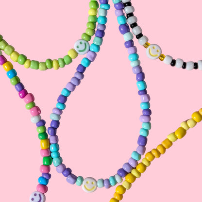 A close-up of several pastel beaded strands arranged on a pink background. The beads come in green, yellow, purple, blue, black, white, and pink, with some strands featuring smiley face charms.