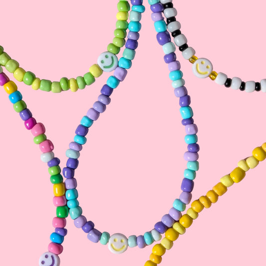 A close-up of several pastel beaded strands arranged on a pink background. The beads come in green, yellow, purple, blue, black, white, and pink, with some strands featuring smiley face charms.