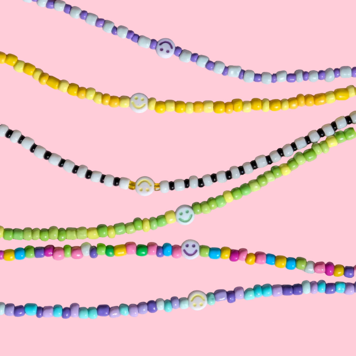 Four strands of colorful beaded necklaces with small smiley face beads, arranged diagonally on a textured light pink background.