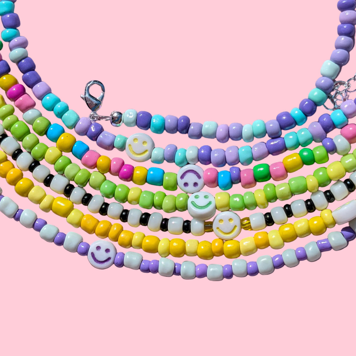 A set of five multi-strand beaded necklaces arranged in a circle on a pink background. The necklaces feature pastel-colored beads in yellow, purple, green, blue, pink, and white, with three white smiley-face beads and silver-tone clasp hardware.