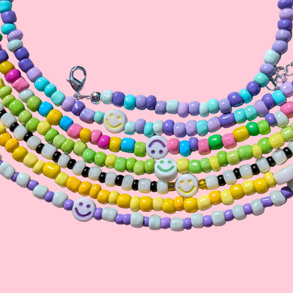 A set of five multi-strand beaded necklaces arranged in a circle on a pink background. The necklaces feature pastel-colored beads in yellow, purple, green, blue, pink, and white, with three white smiley-face beads and silver-tone clasp hardware.