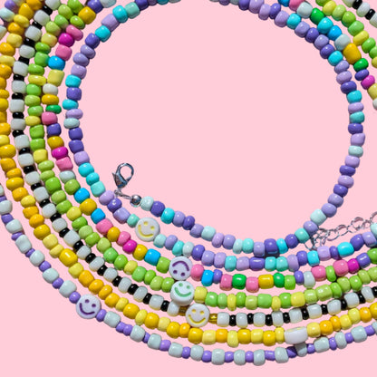 A set of pastel beaded necklaces with smiley face charms, arranged in overlapping loops on a pink background.