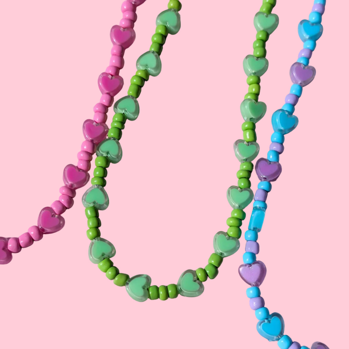 Three strands of heart-shaped beaded necklaces in pink, green, and blue, laid out on a textured light purple background.