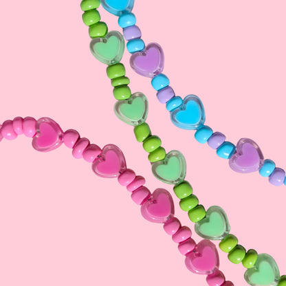 Three strands of heart-shaped beads in pink, green, and blue, arranged on a textured pink background.