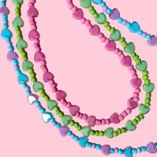 Three strands of heart-shaped beaded necklaces in pink, green, blue, and purple, arranged on a textured pink background.
