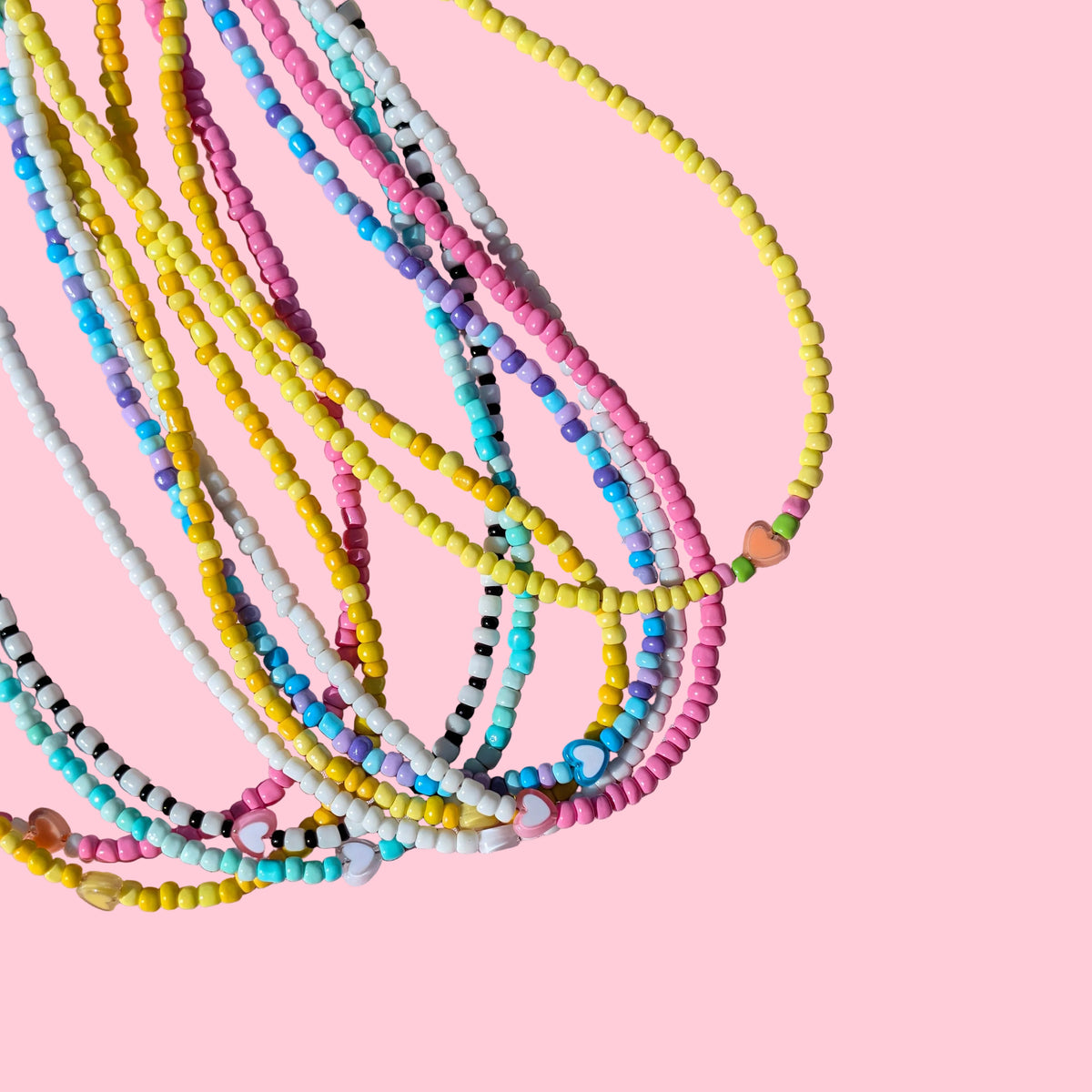 A pile of multi-strand beaded necklaces in pastel colors including yellow, pink, blue, white, and black, arranged on a textured pink background.