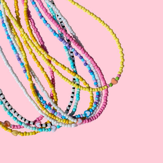 A pile of multi-strand beaded necklaces in pastel colors including yellow, pink, blue, white, and black, arranged on a textured pink background.