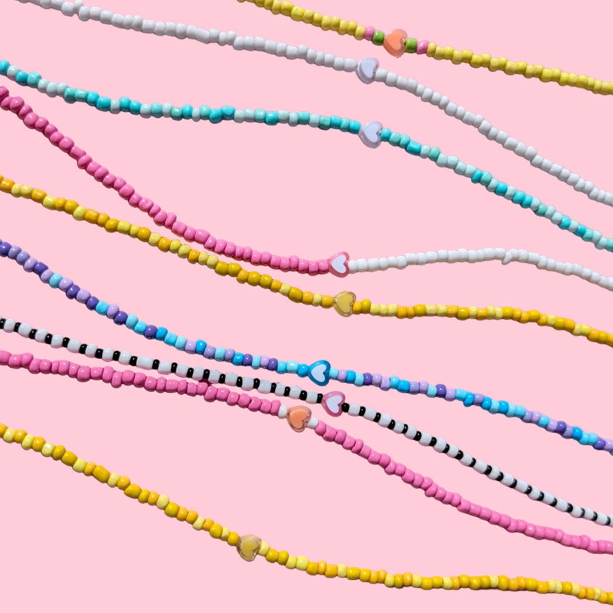 Several strands of small round beads arranged diagonally on a light pink fabric background. The beads come in multiple colorways, including pink, yellow, blue, white, black, and striped patterns.