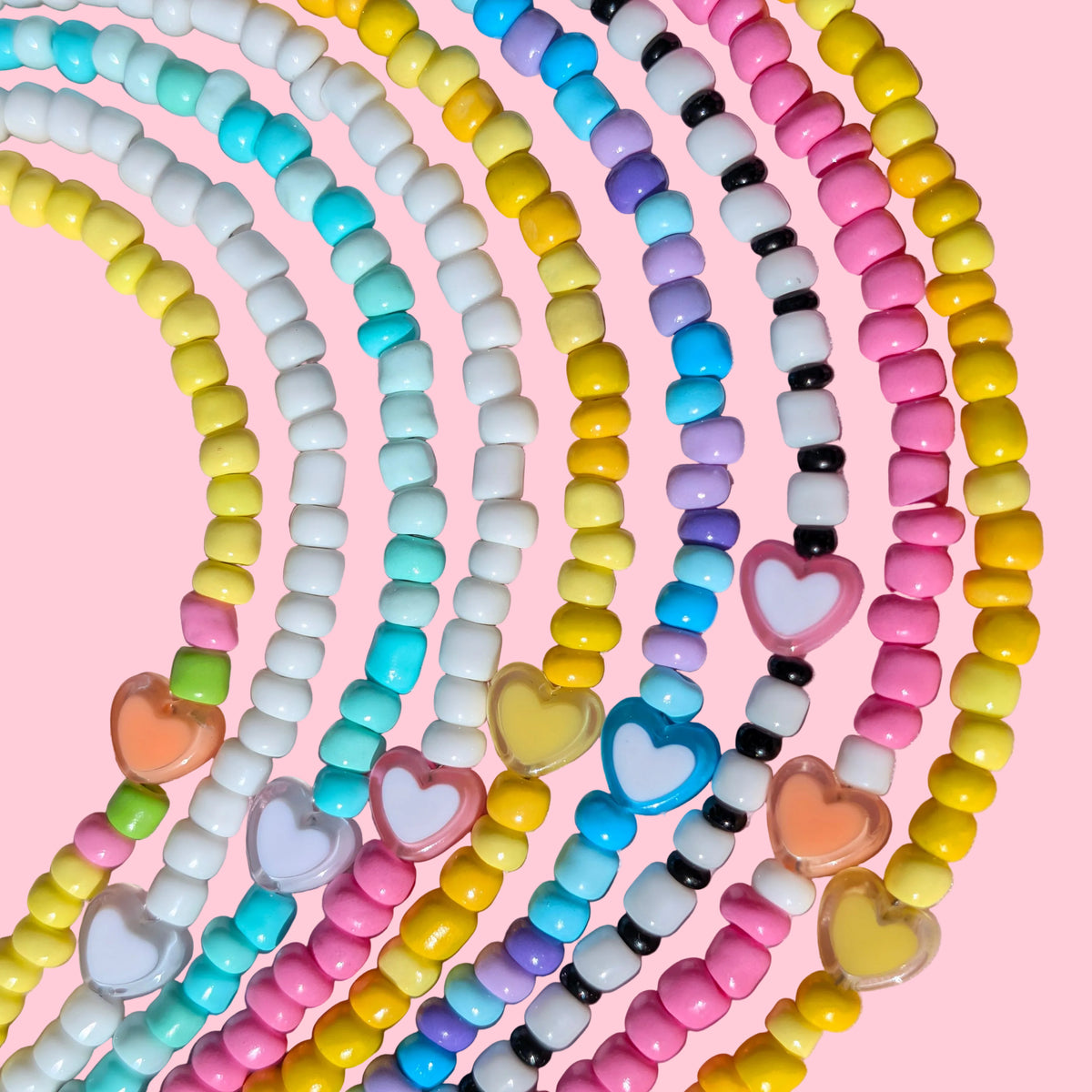 A close-up of multiple pastel beaded necklaces arranged in overlapping curved rows on a pink background. The necklaces feature small round beads in yellow, pink, white, light blue, turquoise, purple, black, and peach, with some translucent heart-shaped beads.