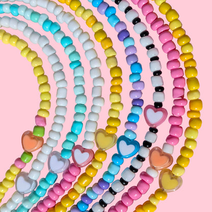 A close-up of multiple pastel beaded necklaces arranged in overlapping curved rows on a pink background. The necklaces feature small round beads in yellow, pink, white, light blue, turquoise, purple, black, and peach, with some translucent heart-shaped beads.