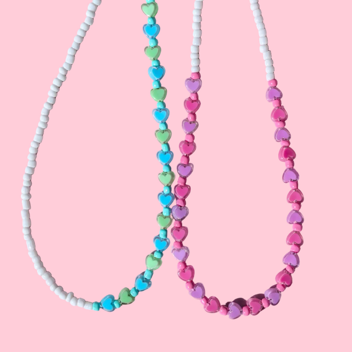 Two beaded necklaces laid flat on a pink background. The top necklace has pink and purple beads with white spacer beads and silver chain.