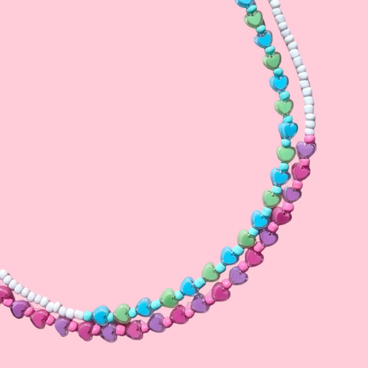 A beaded necklace laid flat on a pink background, featuring alternating strands of pink, blue, green, and white beads with silver-tone metal clasps and extender chains.