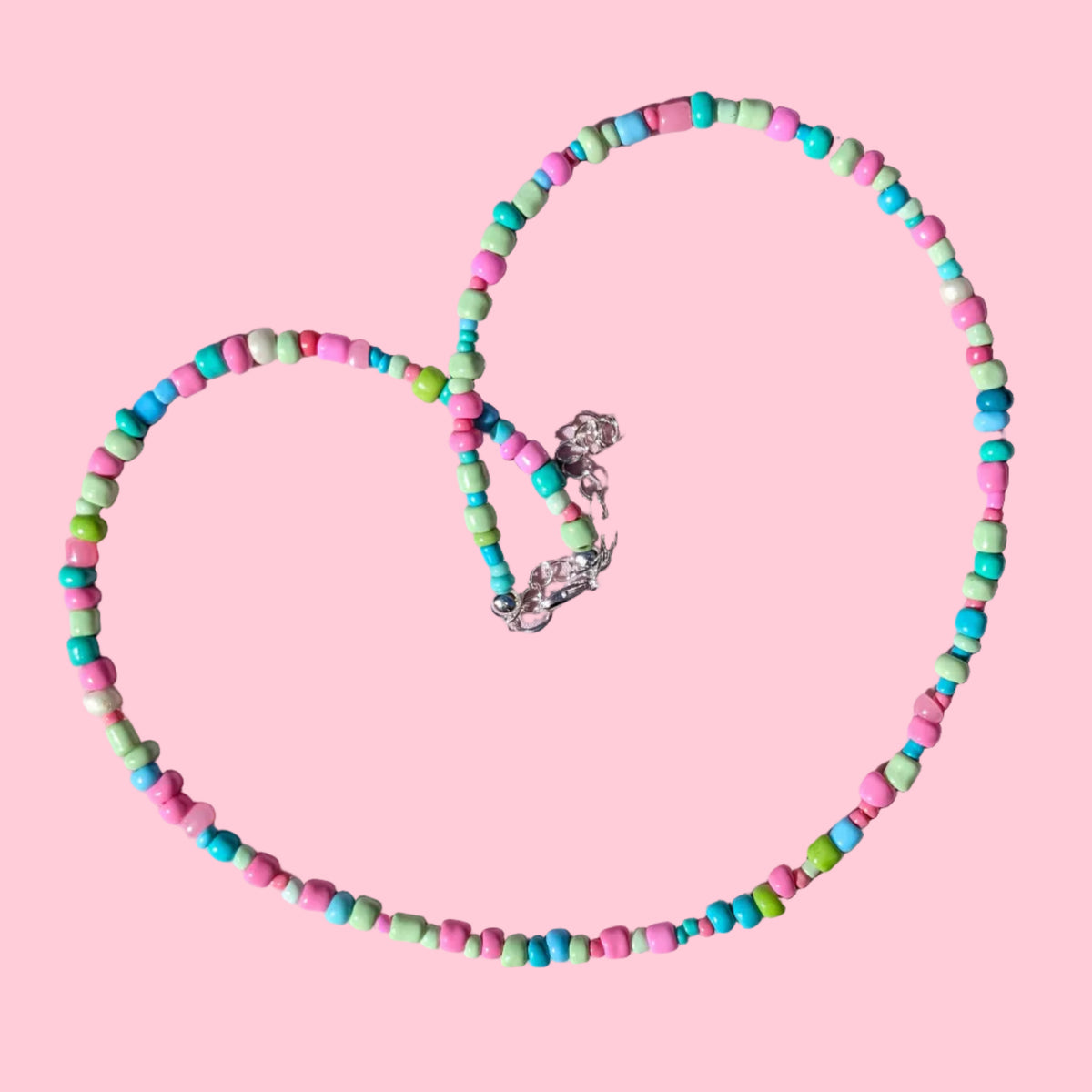 A beaded necklace made of small cylindrical beads in pink, turquoise, and mint green, arranged in a heart shape on a textured light pink background.