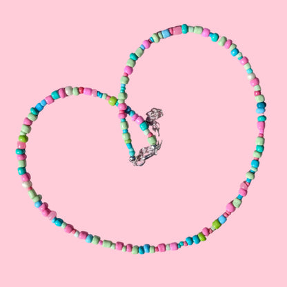 A beaded necklace made of small cylindrical beads in pink, turquoise, and mint green, arranged in a heart shape on a textured light pink background.