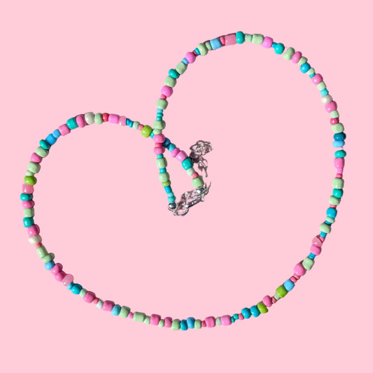 A beaded necklace made of small cylindrical beads in pink, turquoise, and mint green, arranged in a heart shape on a textured light pink background.