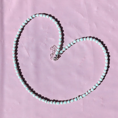 A heart-shaped beaded necklace made of mint green and white beads with a silver-toned heart charm, laid flat on a textured pink background.