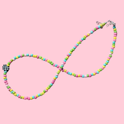 A beaded infinity-style necklace arranged in an infinity shape on a light pink background. The necklace features small cylindrical beads in neon pink, yellow, green, and blue, with a small black-and-white checkered bead near the lower left and a silver clasp at the upper right.