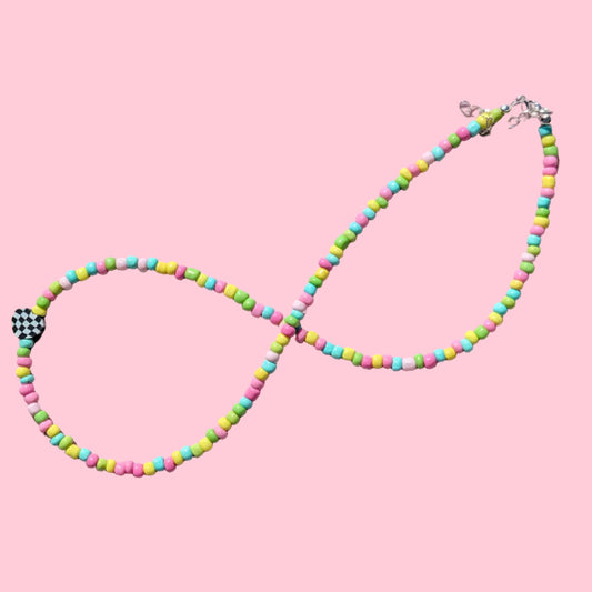 A beaded infinity-style necklace arranged in an infinity shape on a light pink background. The necklace features small cylindrical beads in neon pink, yellow, green, and blue, with a small black-and-white checkered bead near the lower left and a silver clasp at the upper right.