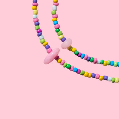 A pastel beaded necklace with small colorful beads and a large light pink disc pendant, arranged on a pink textured background.