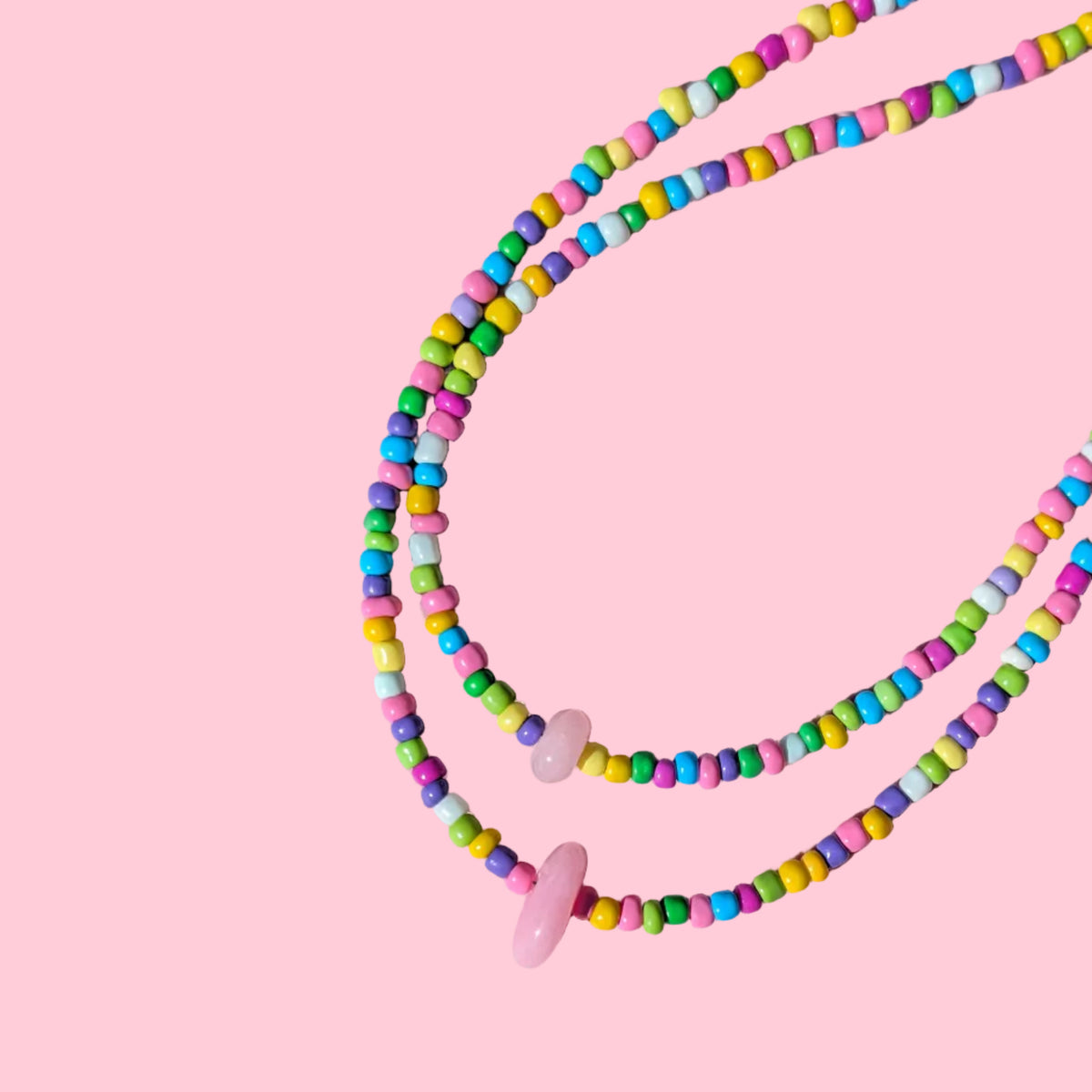 A beaded necklace with small pastel-colored cylindrical beads and two pink oval beads, arranged on a pink background.