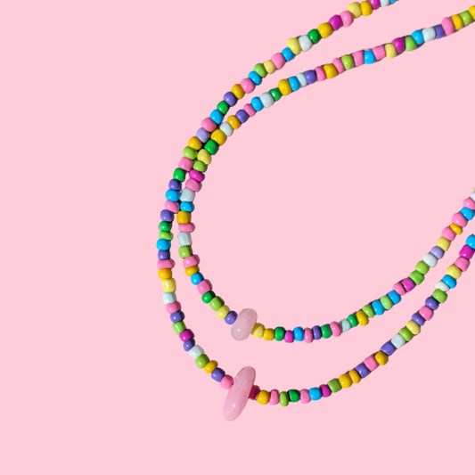 A beaded necklace with small pastel-colored cylindrical beads and two pink oval beads, arranged on a pink background.